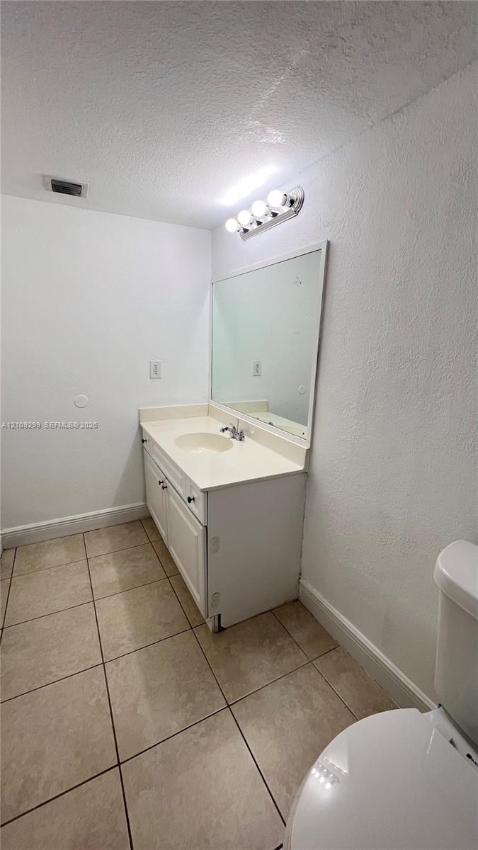 Bathroom featuring a white vanity cabinet, solid-surface integrated countertop with basin, wall-mounted mirror, four-light vanity fixture, and tiled flooring