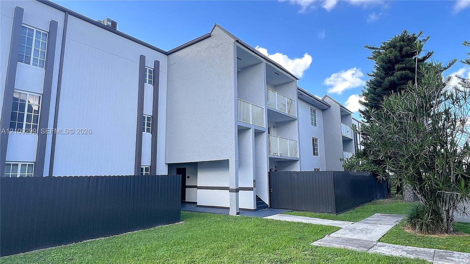Multi-story exterior featuring a light-toned stucco faade, private elevated balconies with metal railings, and dark-finished metal privacy fencing