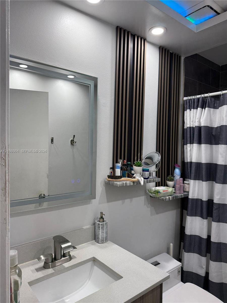 Bathroom featuring a vanity with a stone-surface countertop, an integrated rectangular sink, a polished metal faucet, a backlit rectangular mirror, and decorative wall-mounted acoustic-style slats with built-in stone-finish shelving