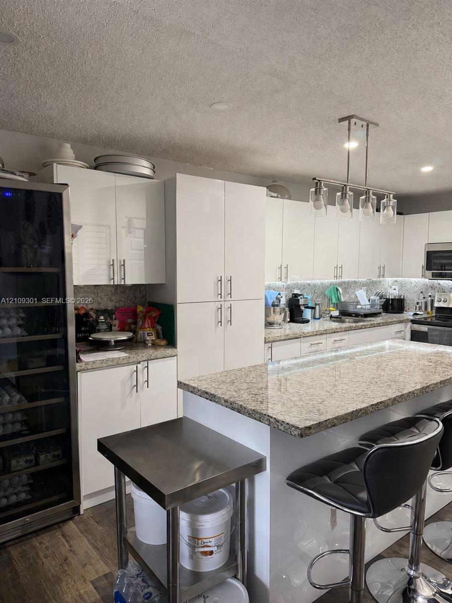 Kitchen featuring glossy white cabinetry, stone countertops, a central island with an integrated breakfast bar, a built-in wine refrigerator, and a linear multi-light pendant fixture
