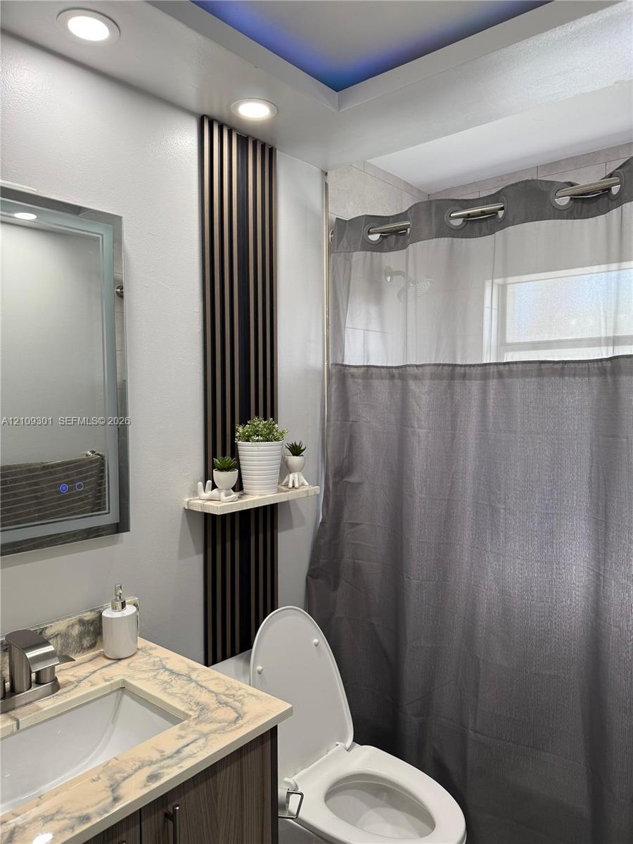 Bathroom featuring a stone-look countertop with an integrated sink, vertical slat accent wall paneling with a floating shelf, recessed ceiling lighting, and a tray ceiling detail with integrated color-accent lighting