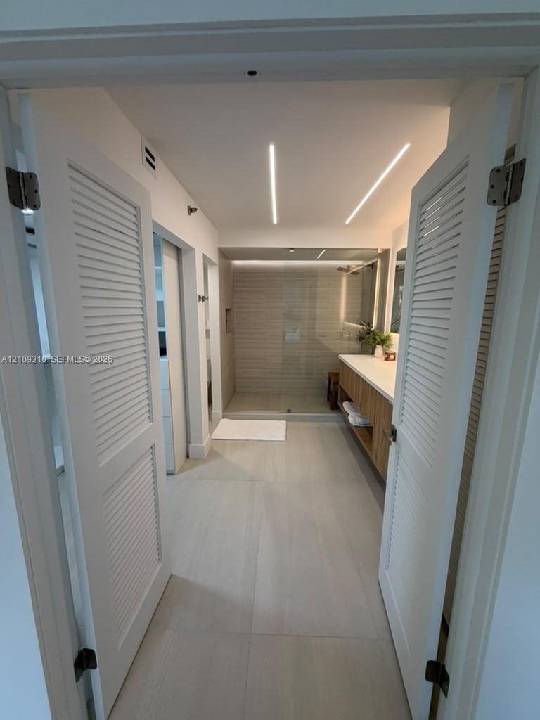 Bathroom featuring dual louvered doors, recessed linear ceiling lighting, floating wood-finish vanity with countertop, and a walk-in glass-enclosed shower with tile surround