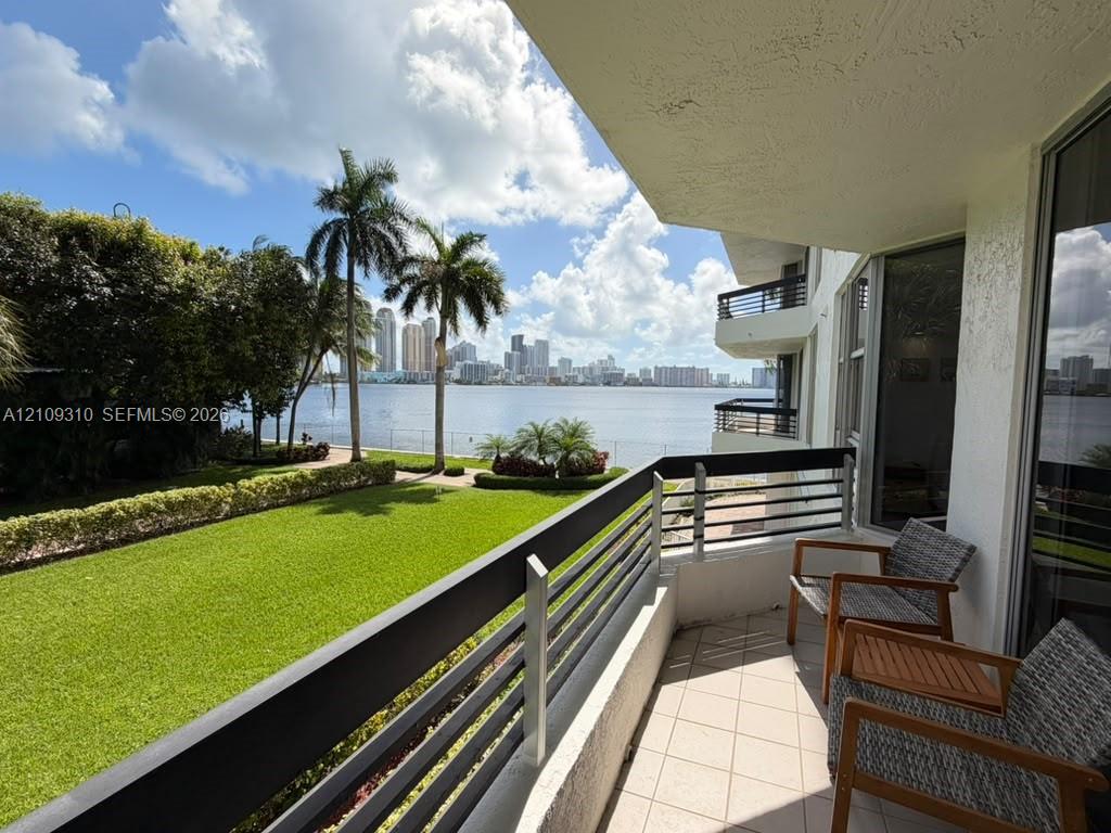 Covered balcony featuring tile flooring and horizontal metal railings, overlooking a waterfront lawn, palm trees, and the city skyline