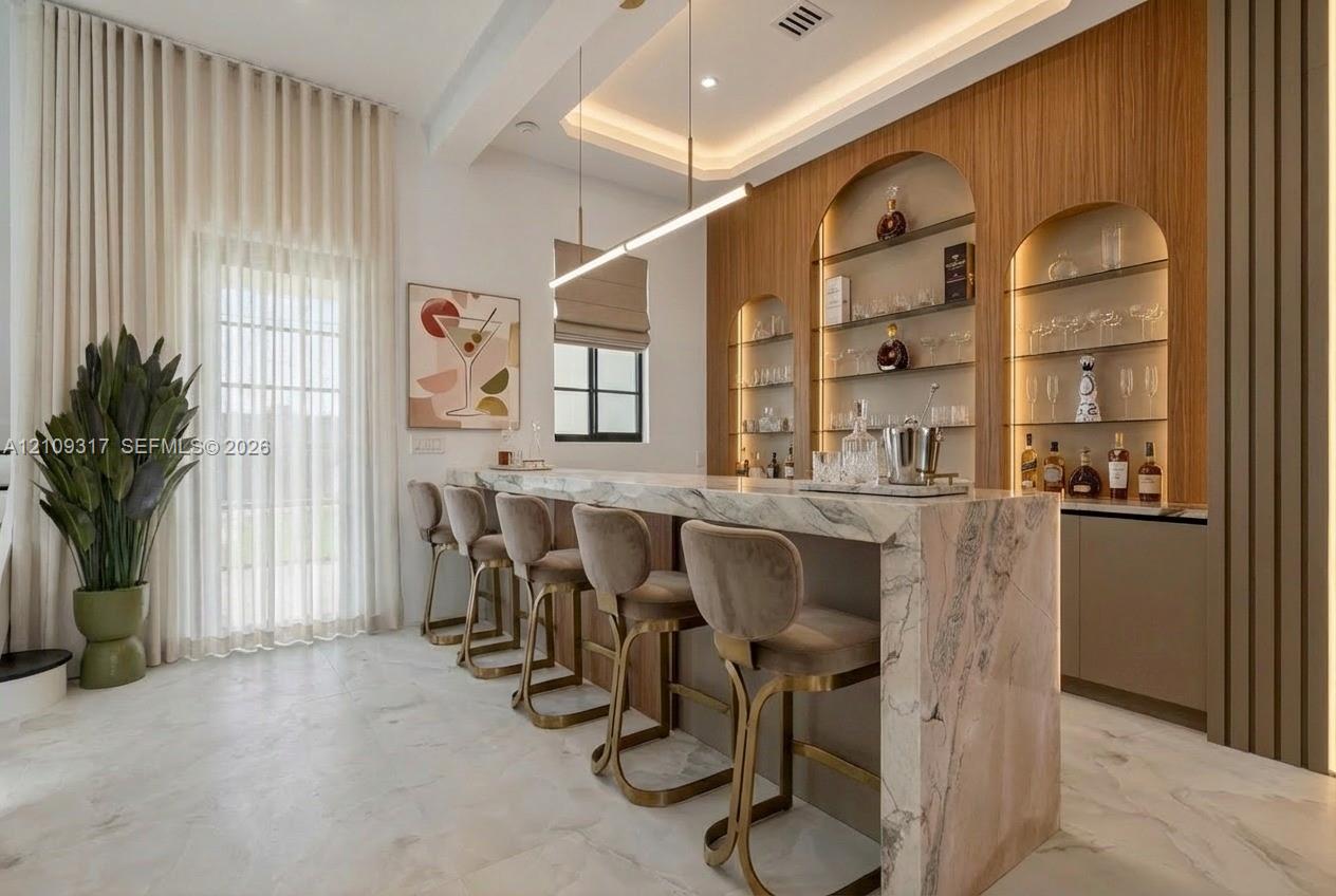 Custom bar area featuring stone countertop and waterfall edge, arched built-in shelving with integrated lighting, wood-finish wall paneling, and tray ceiling with recessed lighting