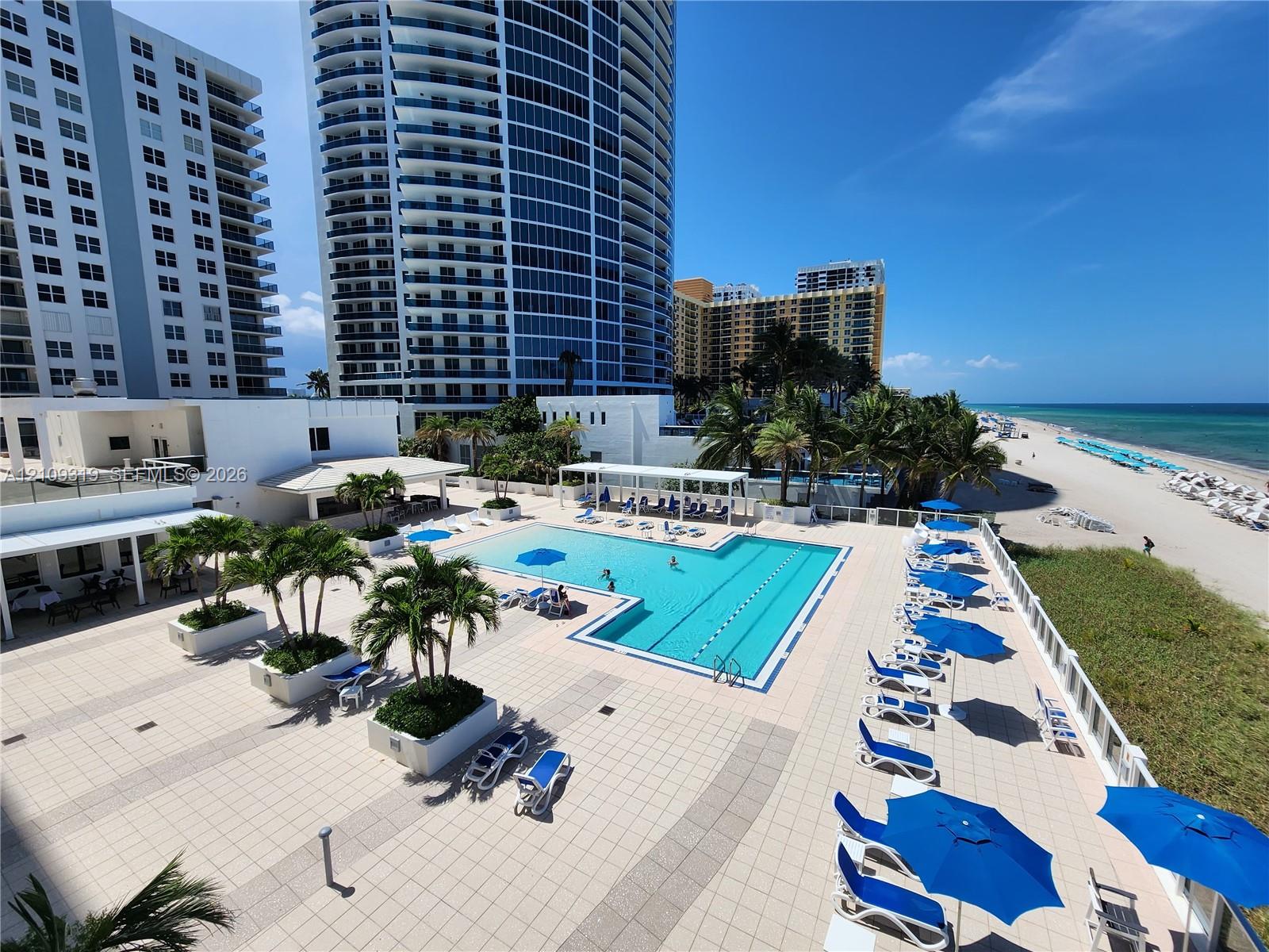 Expansive paved pool deck featuring a swimming pool, integrated landscaping with palms, and direct beachfront access