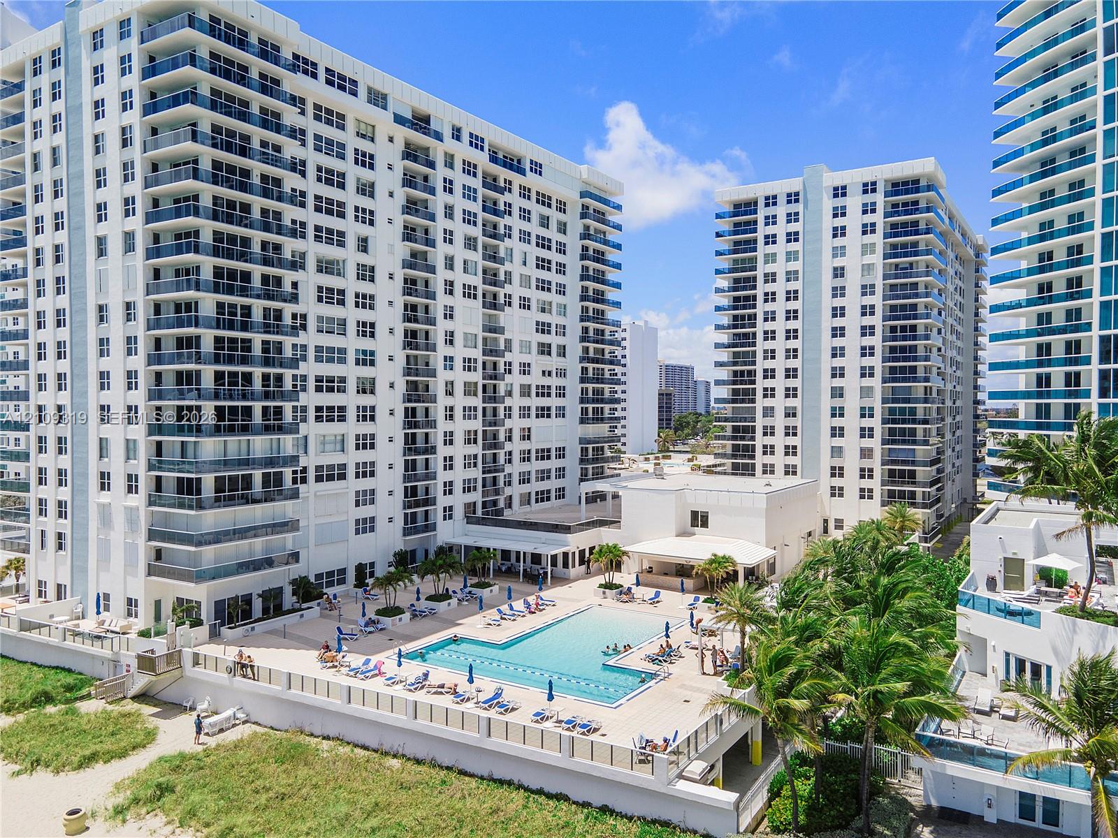 High-rise residential complex featuring a swimming pool, tiled sundeck, palm tree landscaping, and direct access to sandy beach terrain