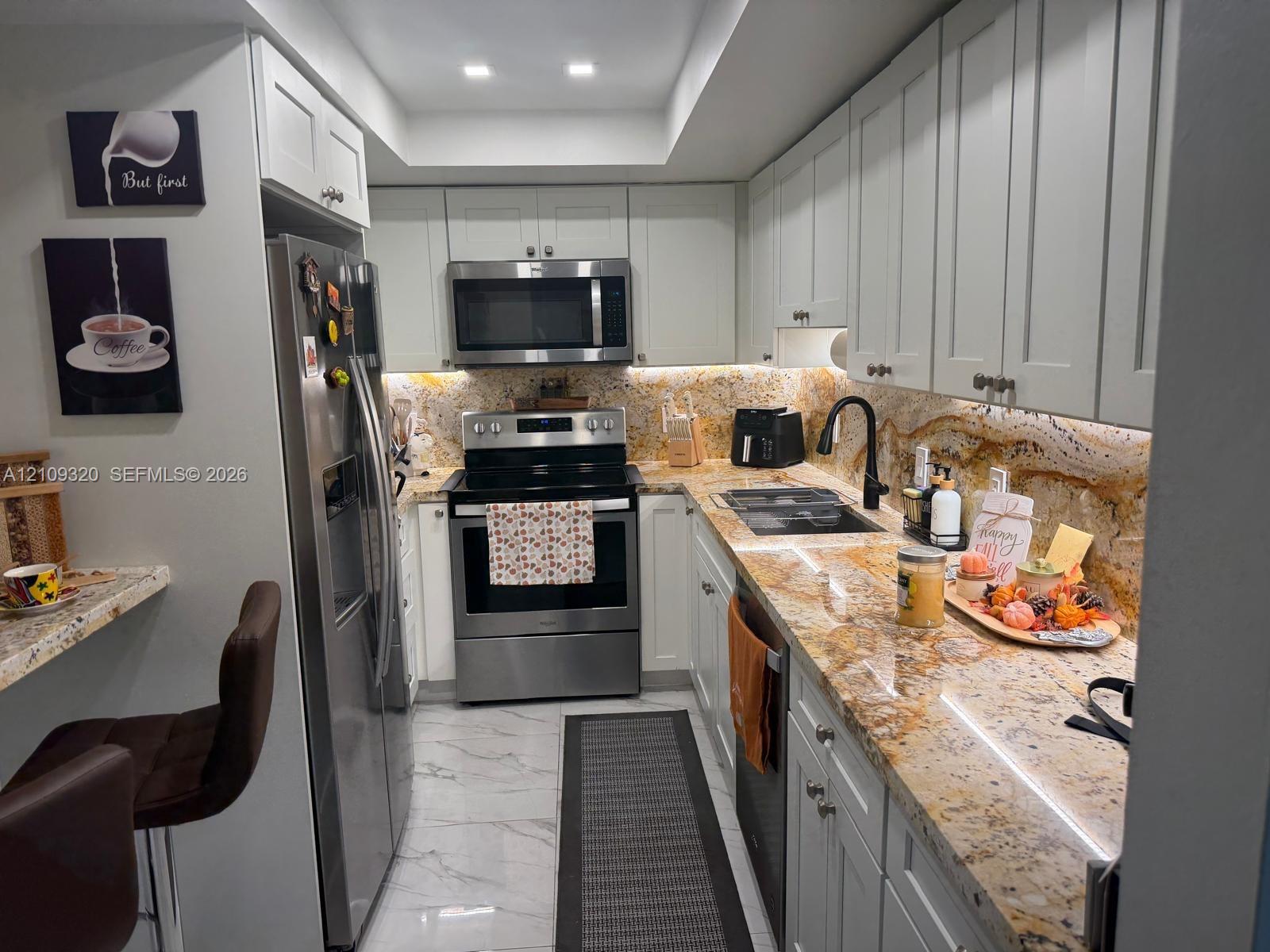 Kitchen featuring light-toned shaker cabinetry, stone countertops with an integrated backsplash, stainless steel appliances, and large-format tile flooring