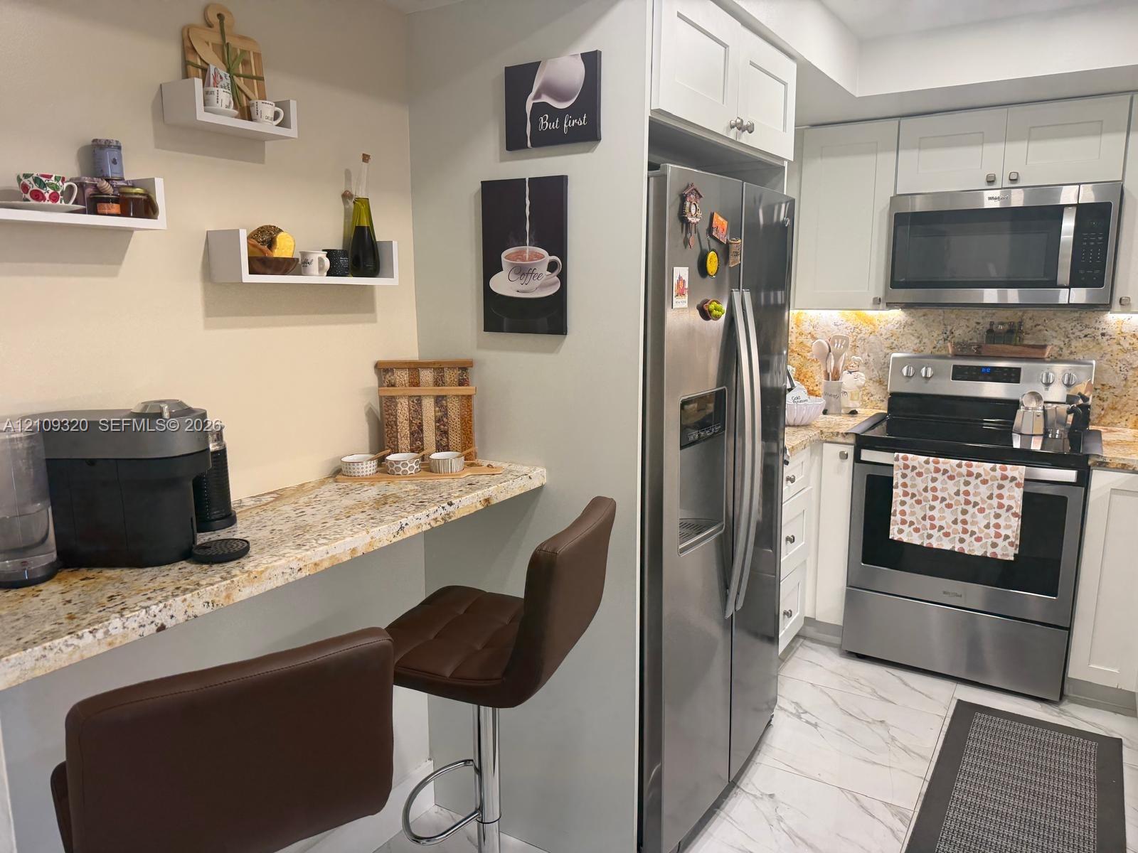 Kitchen featuring light-toned cabinetry, stone-finish countertops, stainless steel appliances, tiled flooring, and wall-mounted floating shelves