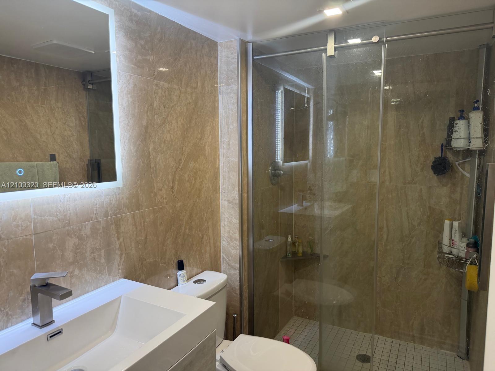 Full-height wall tiling featuring natural stone-patterned surfaces, a wall-mounted LED-backlit mirror, a white rectangular vessel sink with a single-handle chrome faucet, and a glass-enclosed shower with tiled flooring