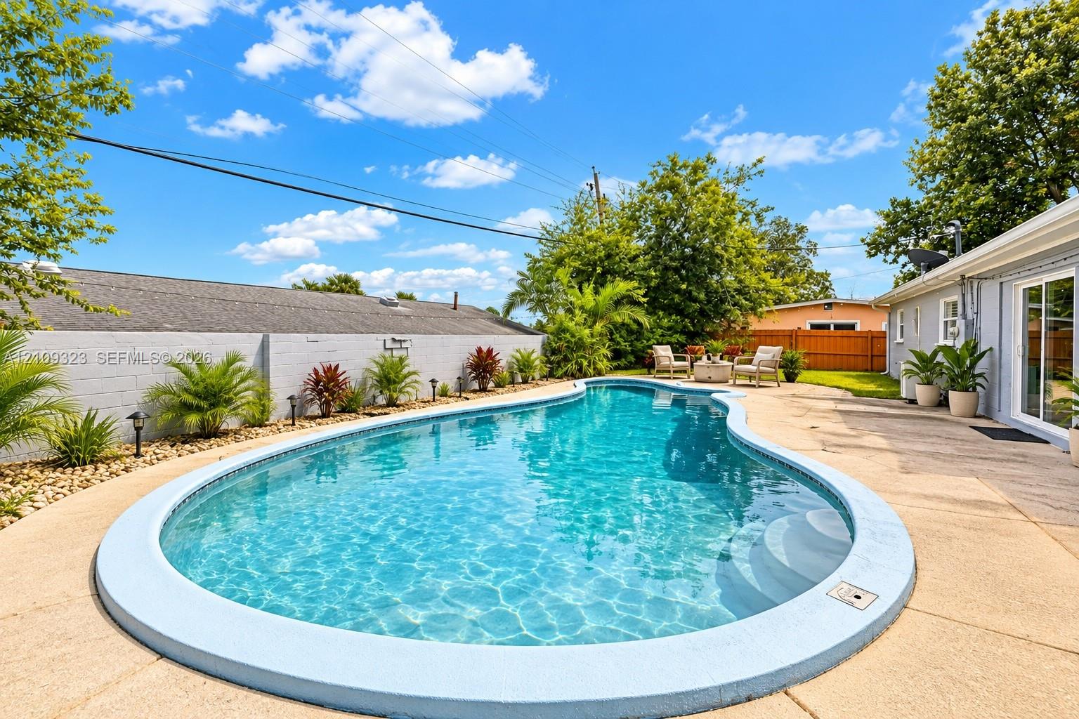 Curved swimming pool featuring a concrete deck, perimeter landscaping, and a masonry privacy wall