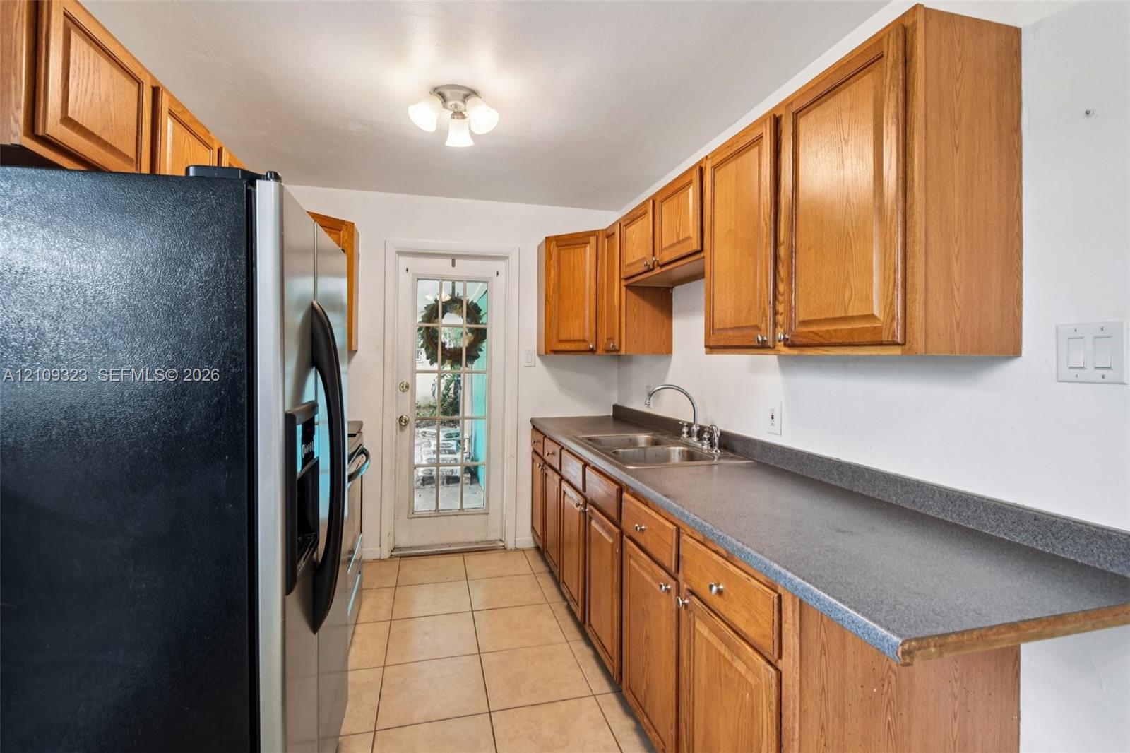 Kitchen featuring wood cabinetry, solid-surface countertops, stainless steel refrigerator, dual-basin sink with faucet, and tile flooring