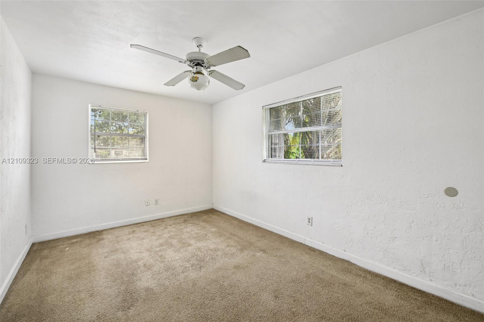 Interior space featuring neutral wall-to-wall carpeting, white textured walls, two windows with multi-pane glass, and a ceiling-mounted fan with integrated lighting