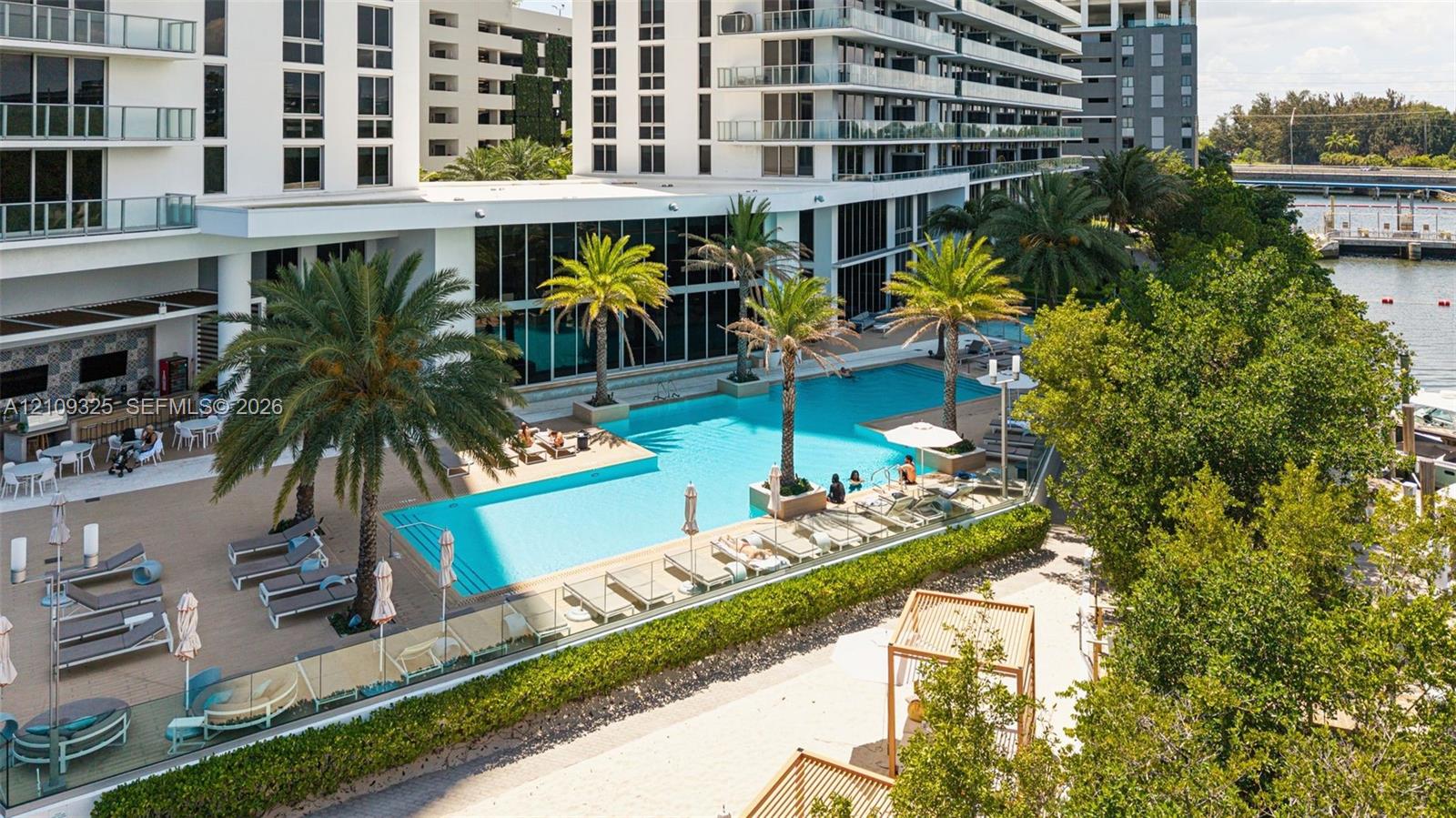 Large swimming pool with palm trees, expansive paved sun deck, glass perimeter fencing, and structured landscaping