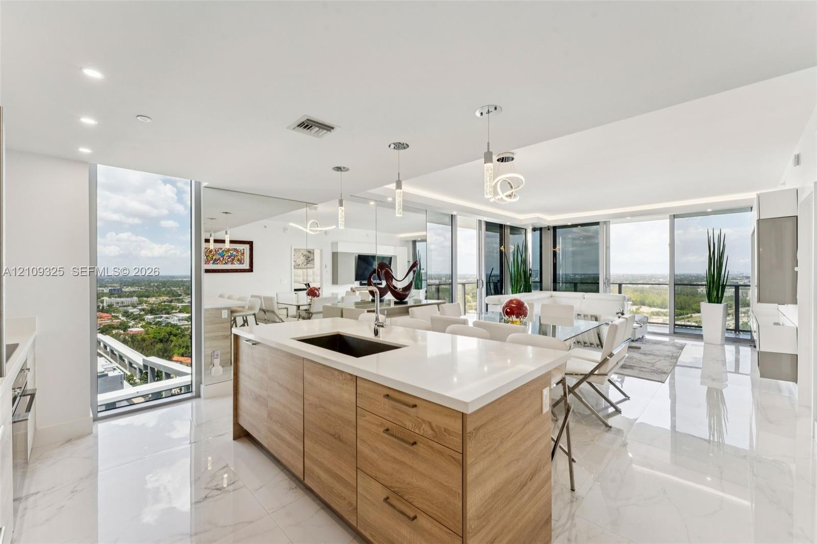 Kitchen island with wood-finish cabinetry, white countertop, and under-mount sink, complemented by polished tile flooring, floor-to-ceiling glass panels, and recessed and pendant lighting