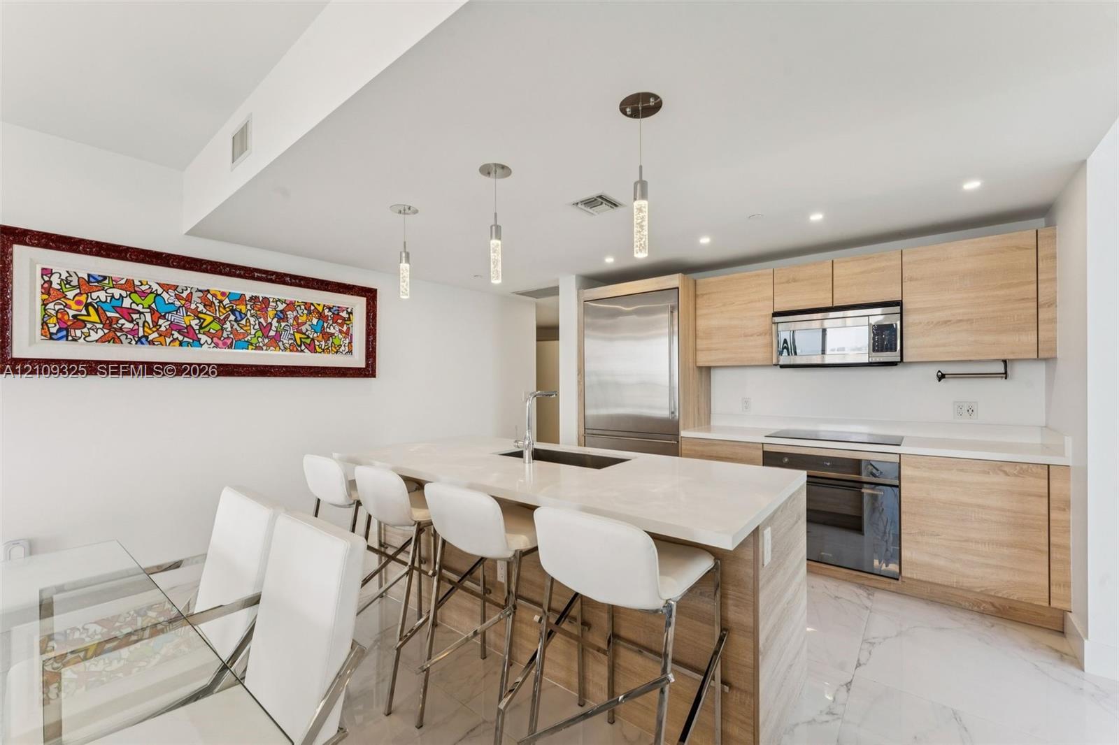 Kitchen featuring wood-finish cabinetry, stone-surface countertops, center island with integrated sink, stainless steel appliances, and glass pendant lighting over marble-finish flooring