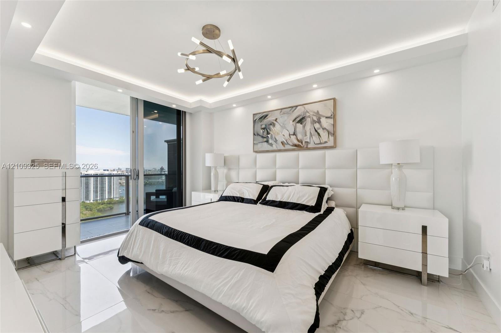 White stone-finish flooring with grey veining, recessed tray ceiling with LED cove lighting, modern tiered metallic chandelier, floor-to-ceiling sliding glass doors, and built-in upholstered grid-pattern headboard