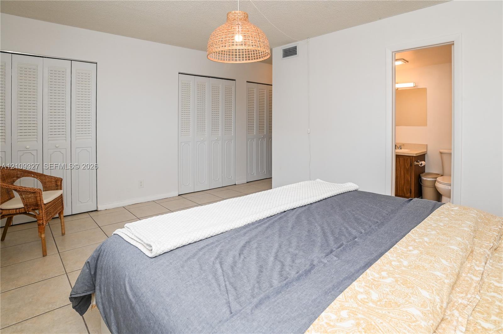 Room featuring neutral tile flooring, multiple sets of white louvered-door closets, a woven pendant light fixture, and an entry to an en-suite bathroom