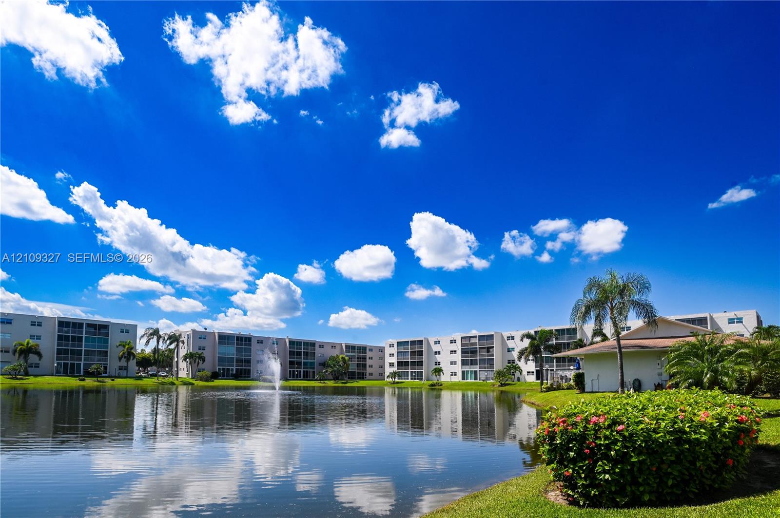 Waterfront community grounds featuring a central fountain, manicured landscaping, palm trees, and residential buildings
