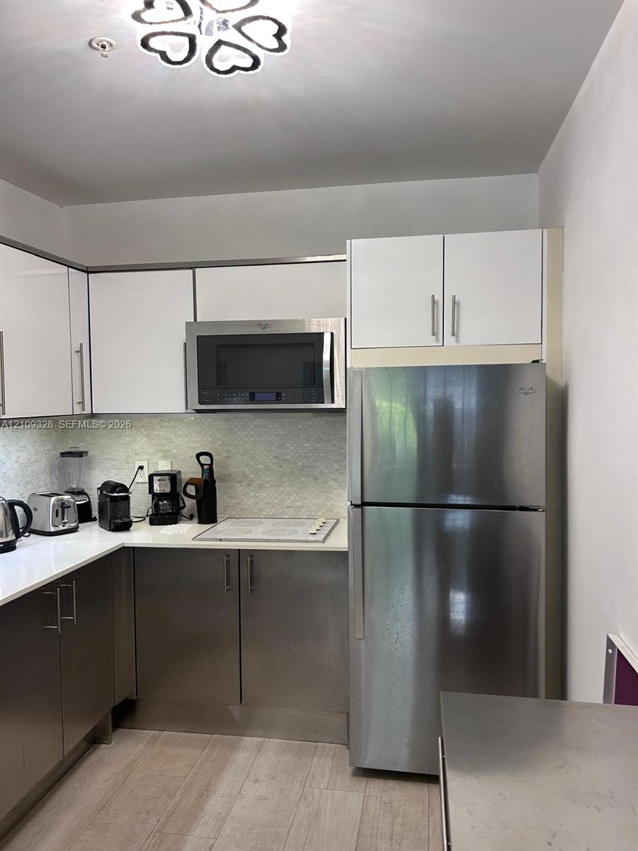 Kitchen featuring two-tone cabinetry with white upper and stainless steel lower doors, stainless steel refrigerator and built-in microwave, solid surface countertops, mosaic tile backsplash, and light-toned wood-finish flooring