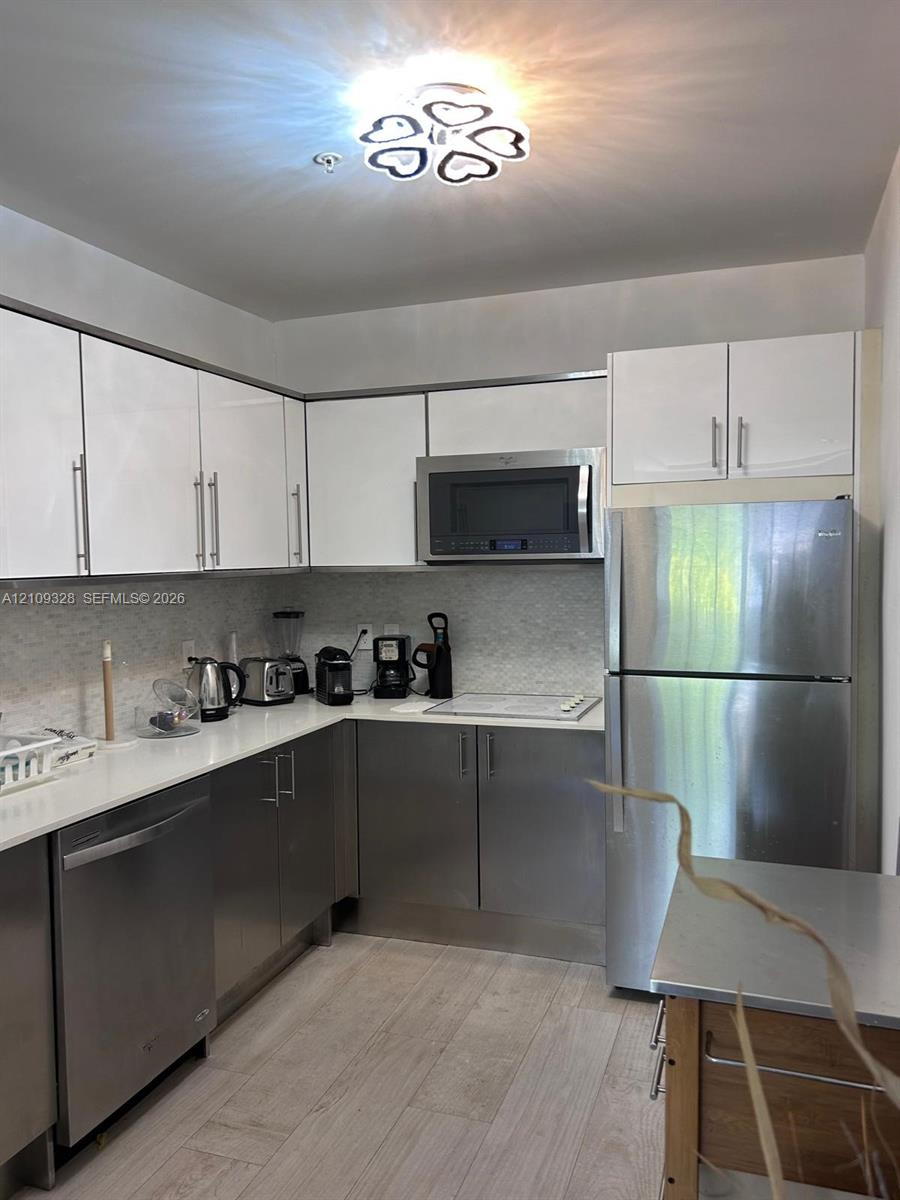 Kitchen featuring white upper cabinetry, stainless steel lower cabinetry, solid-surface countertops, textured tile backsplash, and stainless steel appliances