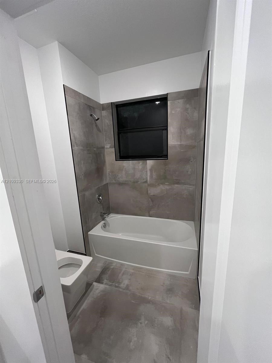 Full-height tiled walls surrounding a soaking tub with integrated shower hardware, large-format tile flooring, and a window with a black frame
