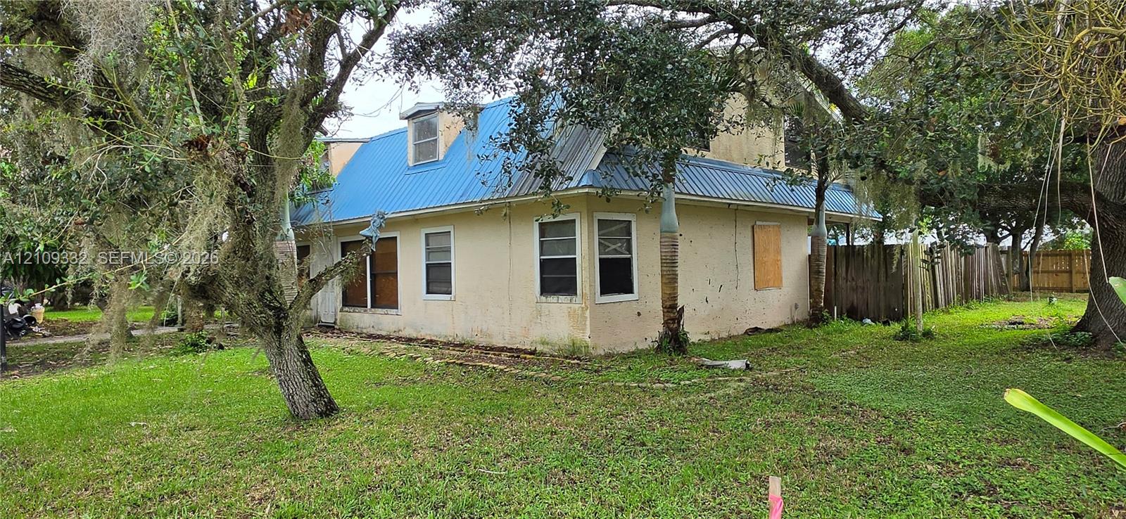 Exterior featuring a light-colored stucco facade, blue standing-seam metal roofing, dormer window, and mature trees on the surrounding grounds