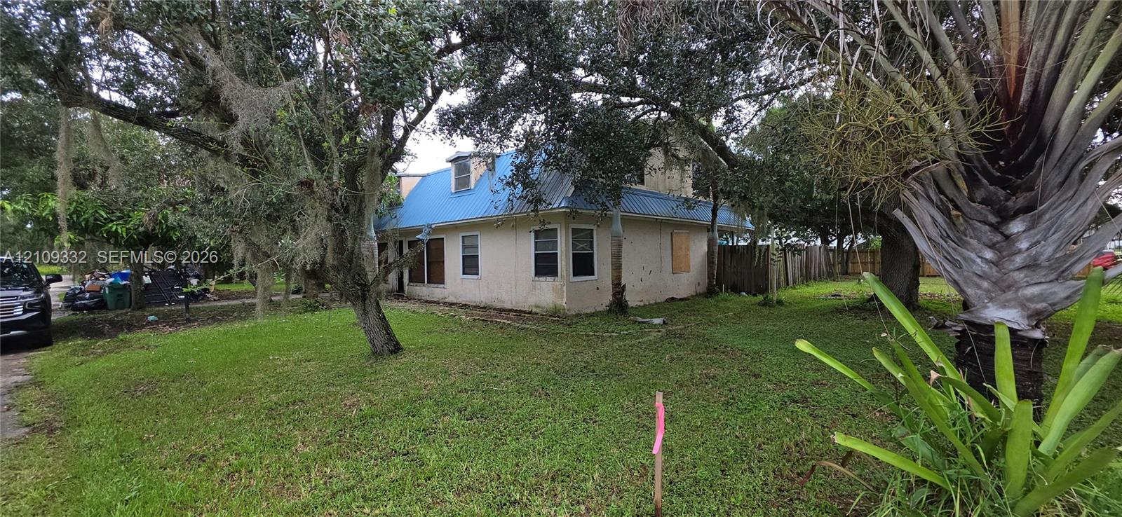 Exterior featuring a light-colored stucco finish, a blue metal roof, multiple windows with grid detailing, a dormer window, and mature oak trees on the property