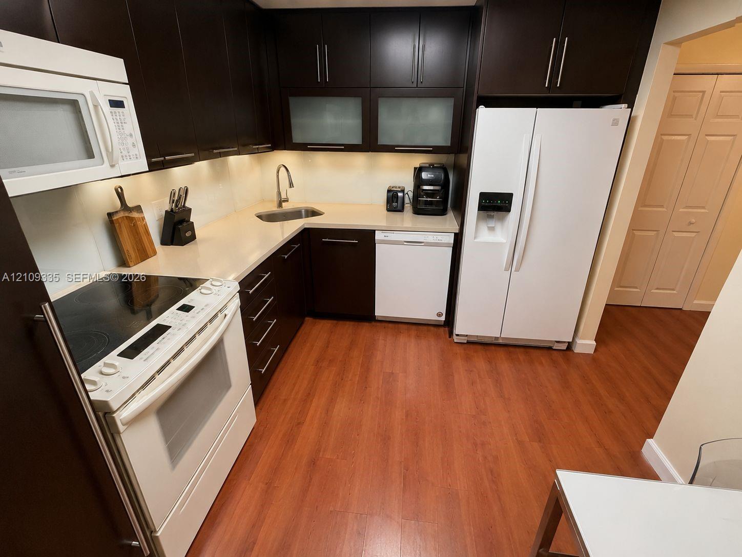 Kitchen featuring dark wood-finish cabinetry, solid surface countertops, glass-paneled upper cabinets, and wood-finish flooring Kitchen featuring dark wood-finish cabinetry, solid surface countertops, glass-paneled upper cabinets, and wood-finish flooring
