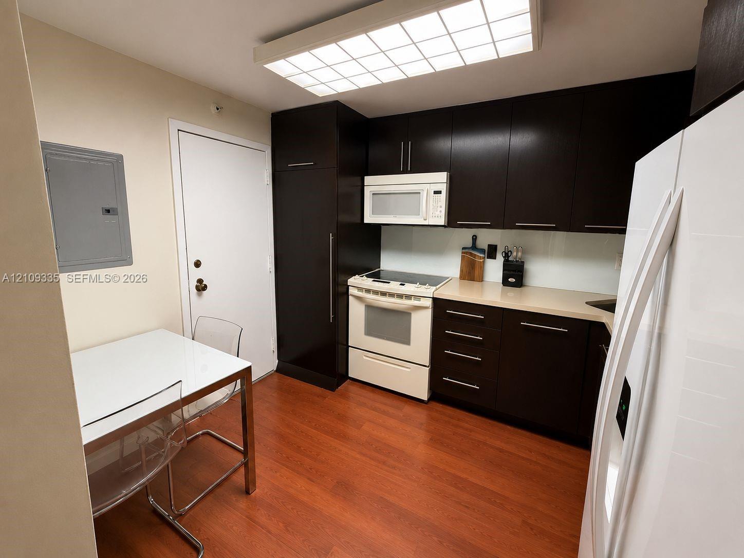 Kitchen featuring dark-finish cabinetry, solid-surface countertops, wood-finish flooring, integrated refrigeration, and a flush-mount grid ceiling light Kitchen featuring dark-finish cabinetry, solid-surface countertops, wood-finish flooring, integrated refrigeration, and a flush-mount grid ceiling light