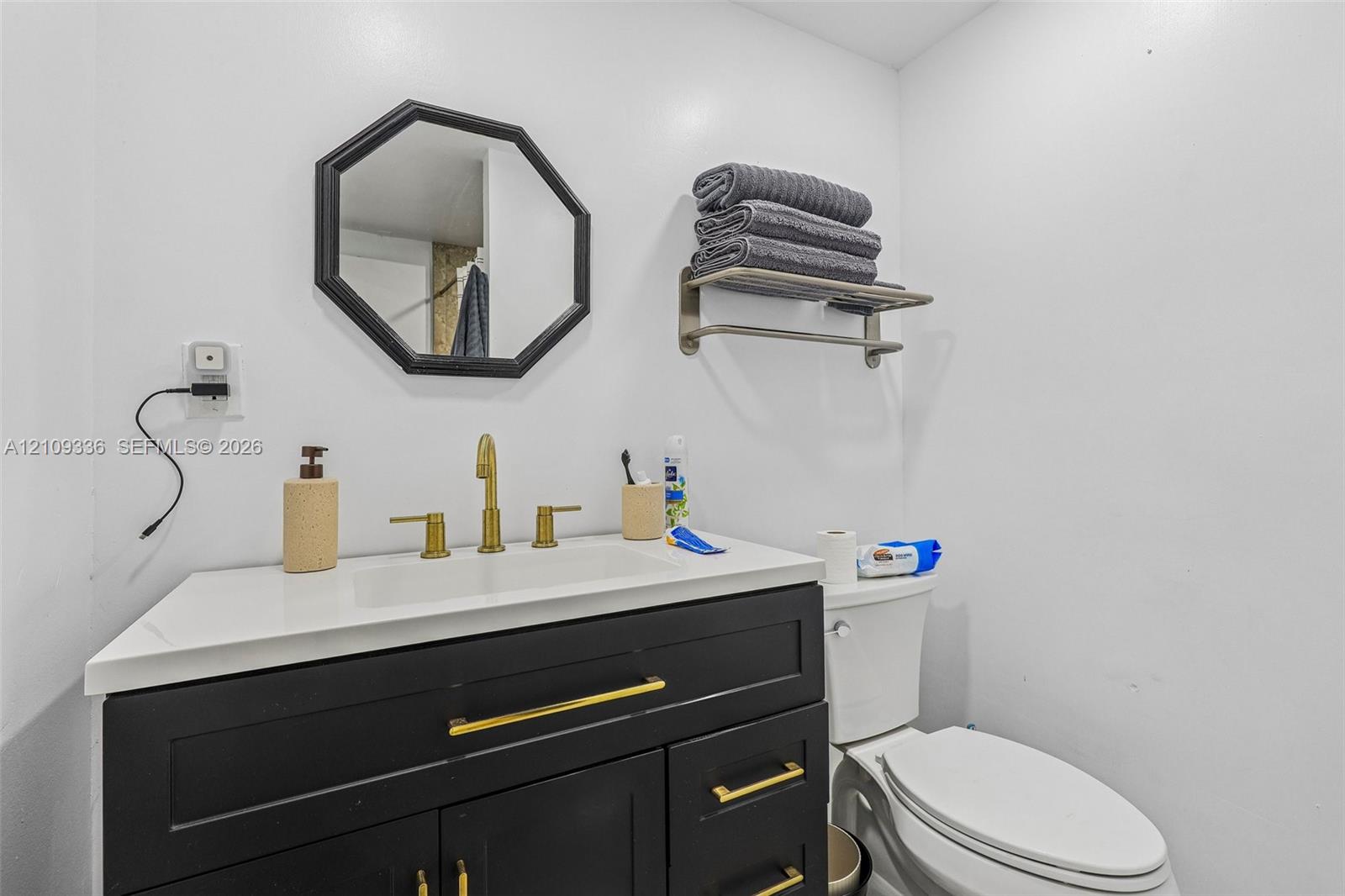 Dark cabinetry vanity with gold-tone hardware, rectangular white countertop basin, brushed gold widespread faucet, wall-mounted octagonal mirror, and metal wall-mounted shelving