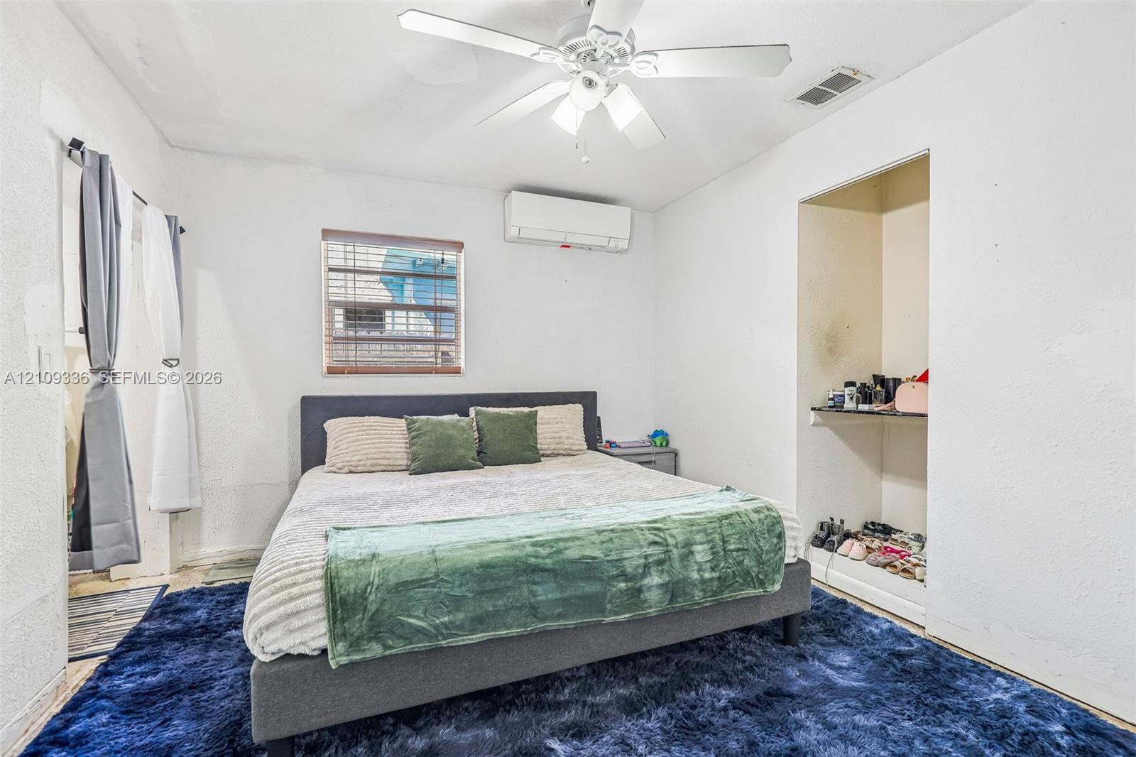 Bedroom featuring a ceiling fan with integrated lighting, a wall-mounted ductless mini-split system, a window with horizontal blinds, and a built-in closet alcove with shelving