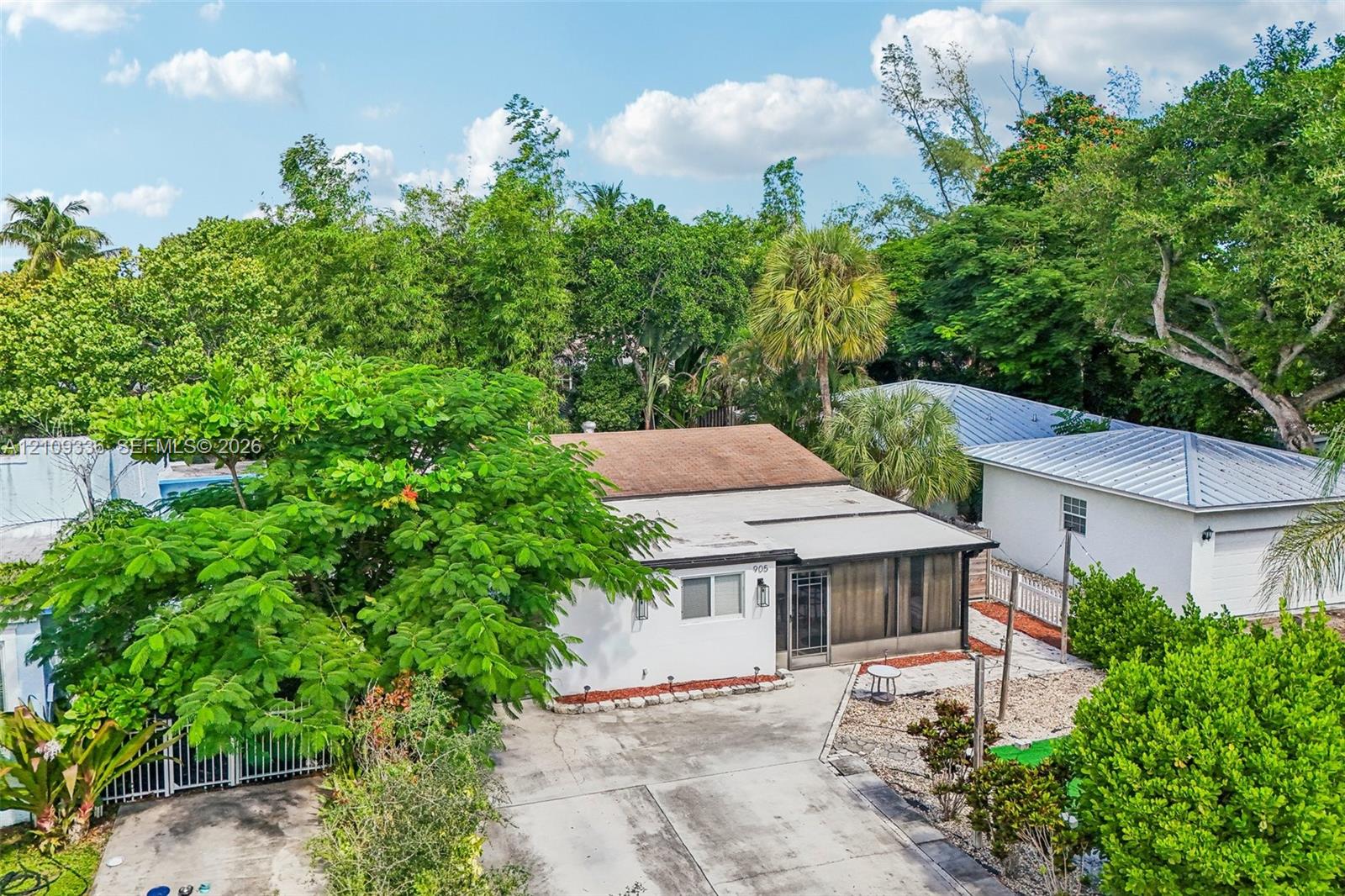 Single-story residence featuring a shingled roof, stucco exterior, concrete driveway, and a screened-in porch, surrounded by mature trees and lush vegetation