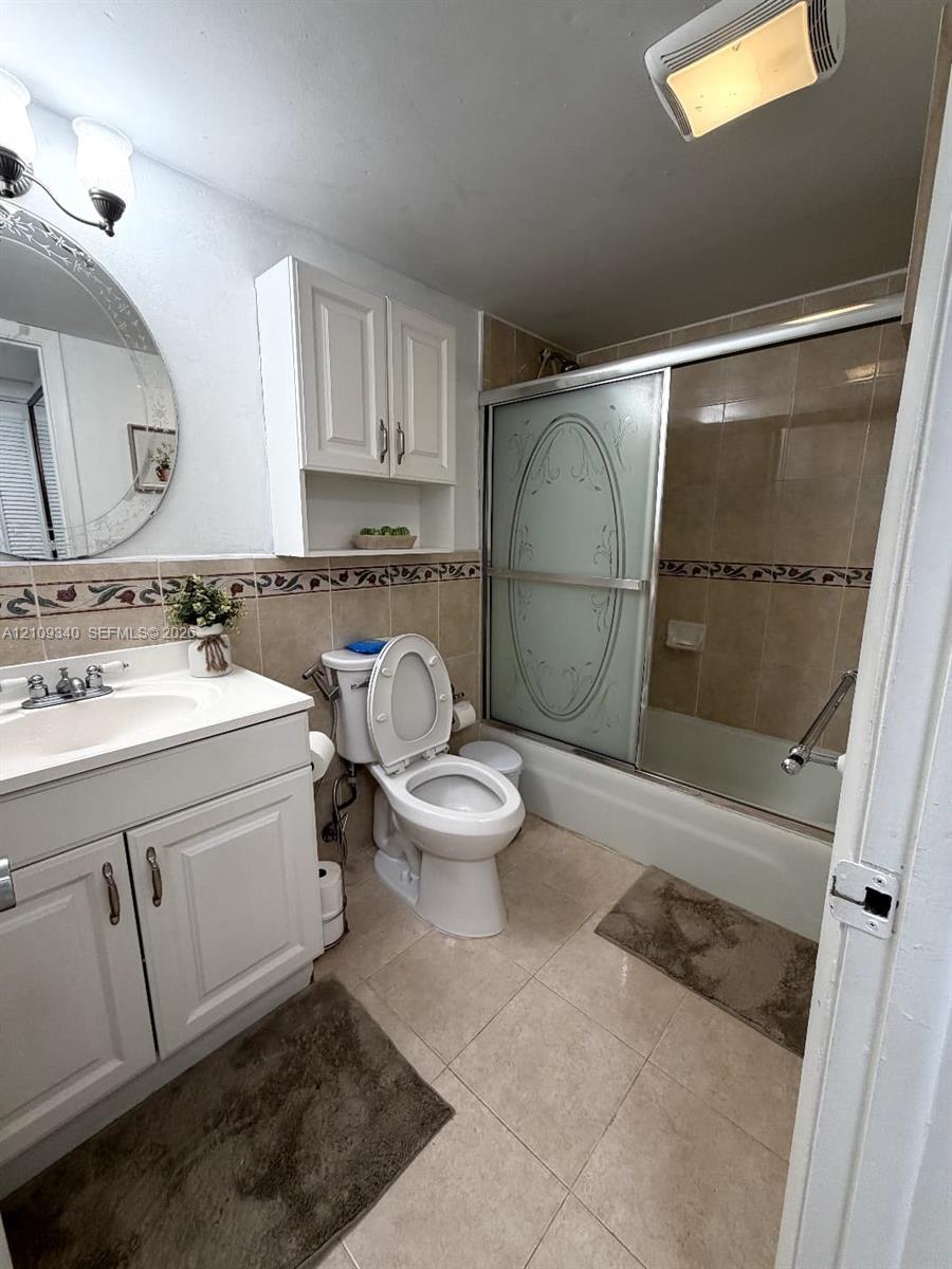Bathroom featuring a white vanity cabinet with an integrated sink, wall-mounted storage cabinetry, a recessed bathtub with a decorative glass sliding door, and tile flooring