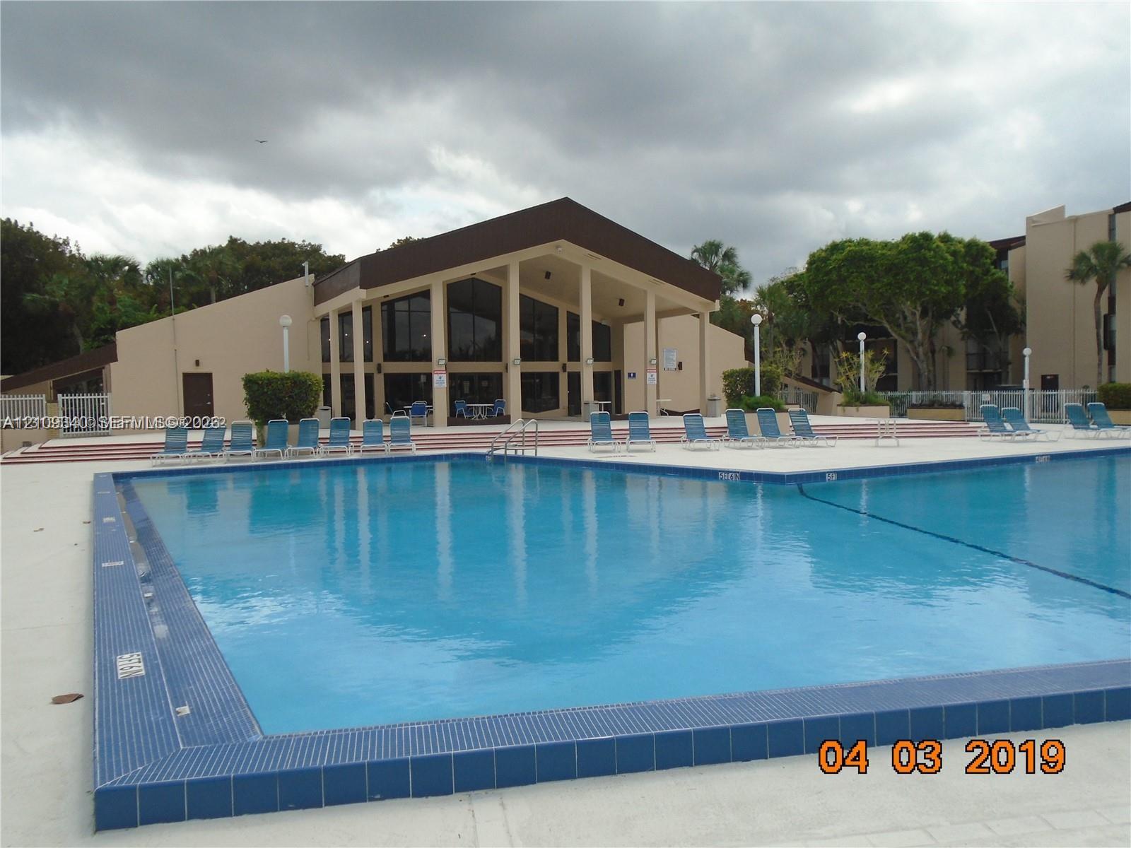In-ground swimming pool with a tiled perimeter, expansive concrete deck, and multi-level structure featuring a gabled roof and floor-to-ceiling windows