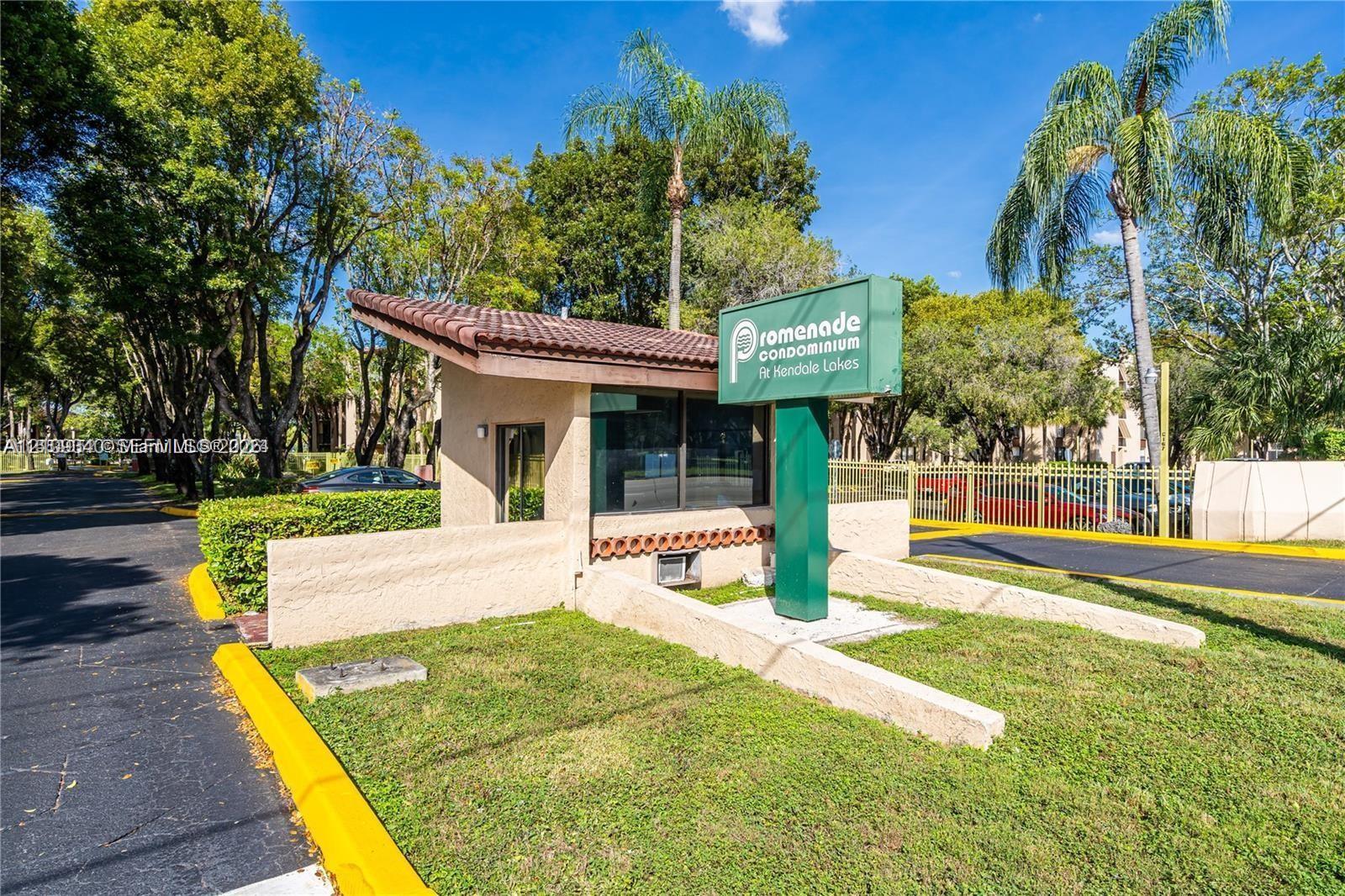 Gated community entrance featuring a guardhouse with a tiled roof, manicured landscaping, and mature palm trees