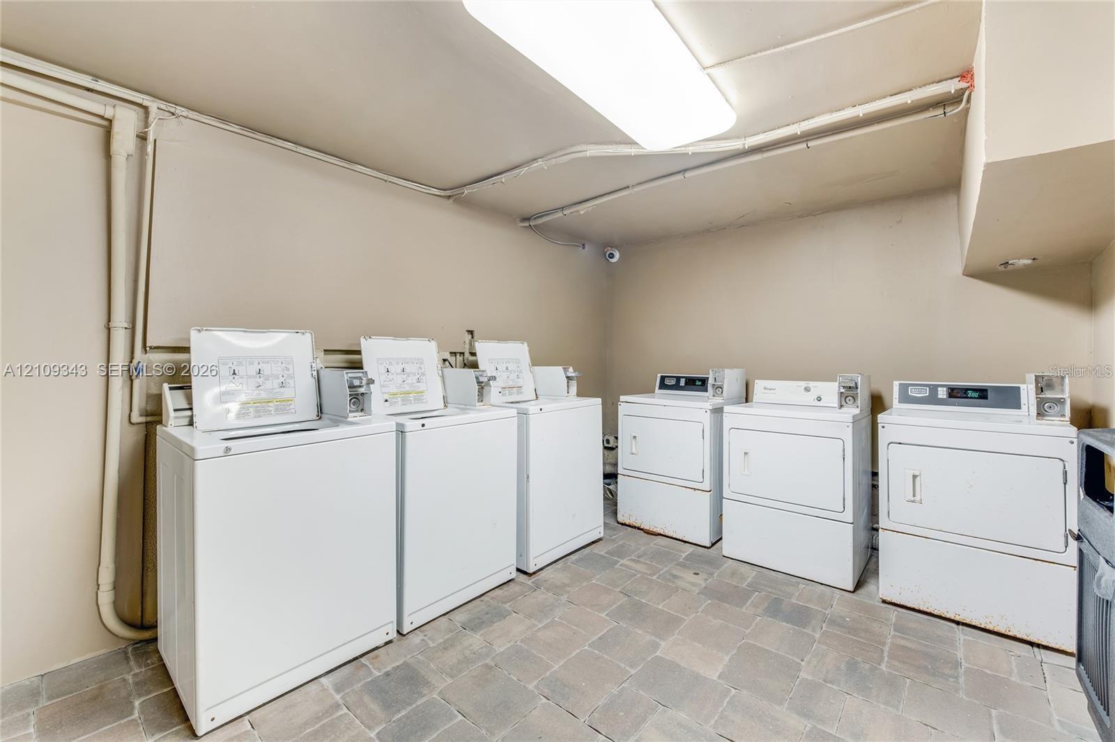 Utility room featuring a multi-unit laundry configuration with three top-load washers and three front-load dryers, complemented by stone-finish tile flooring