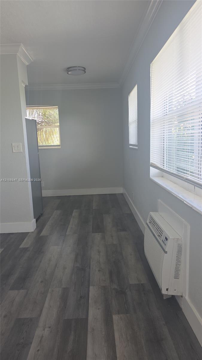 Wood-finish flooring, neutral wall tones, crown molding, windows with integrated blinds, and wall-mounted climate control unit