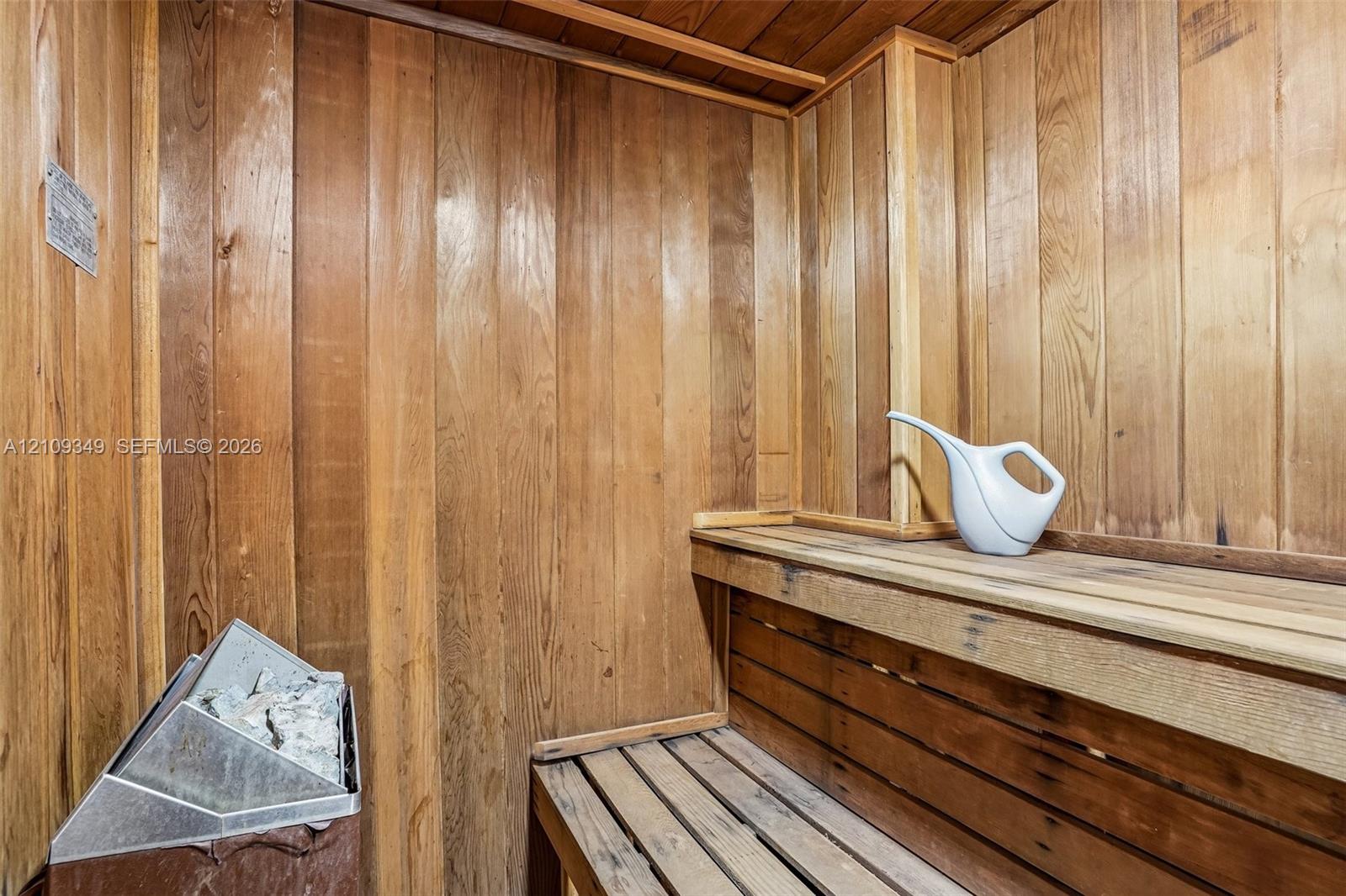 Sauna featuring tongue-and-groove wood wall paneling, multi-level wood bench seating, and a metal heating unit with stones