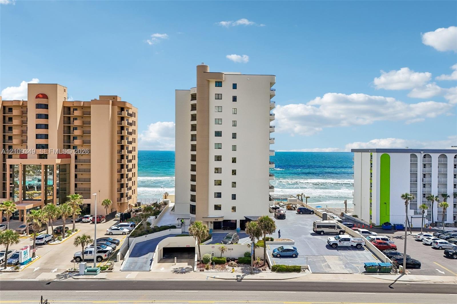 High-rise beachfront building featuring a multi-story exterior, paved parking area, ground-level covered parking access, and mature palm trees High-rise beachfront building featuring a multi-story exterior, paved parking area, ground-level covered parking access, and mature palm trees
