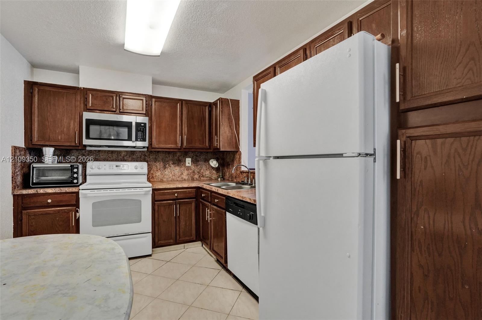 Kitchen featuring dark wood-finish cabinetry, white appliances, stone-patterned countertops, and tile flooring