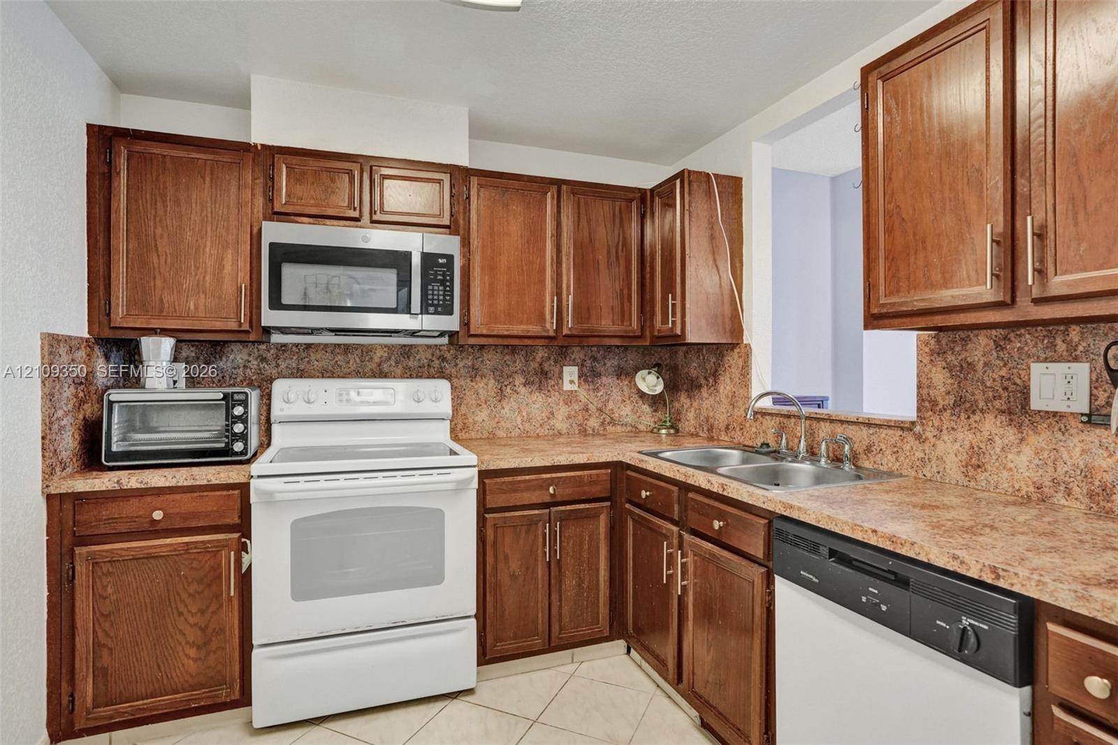 Kitchen featuring wood cabinetry, stone-finish laminate countertops, dual-basin stainless steel sink, tile flooring, and integrated microwave
