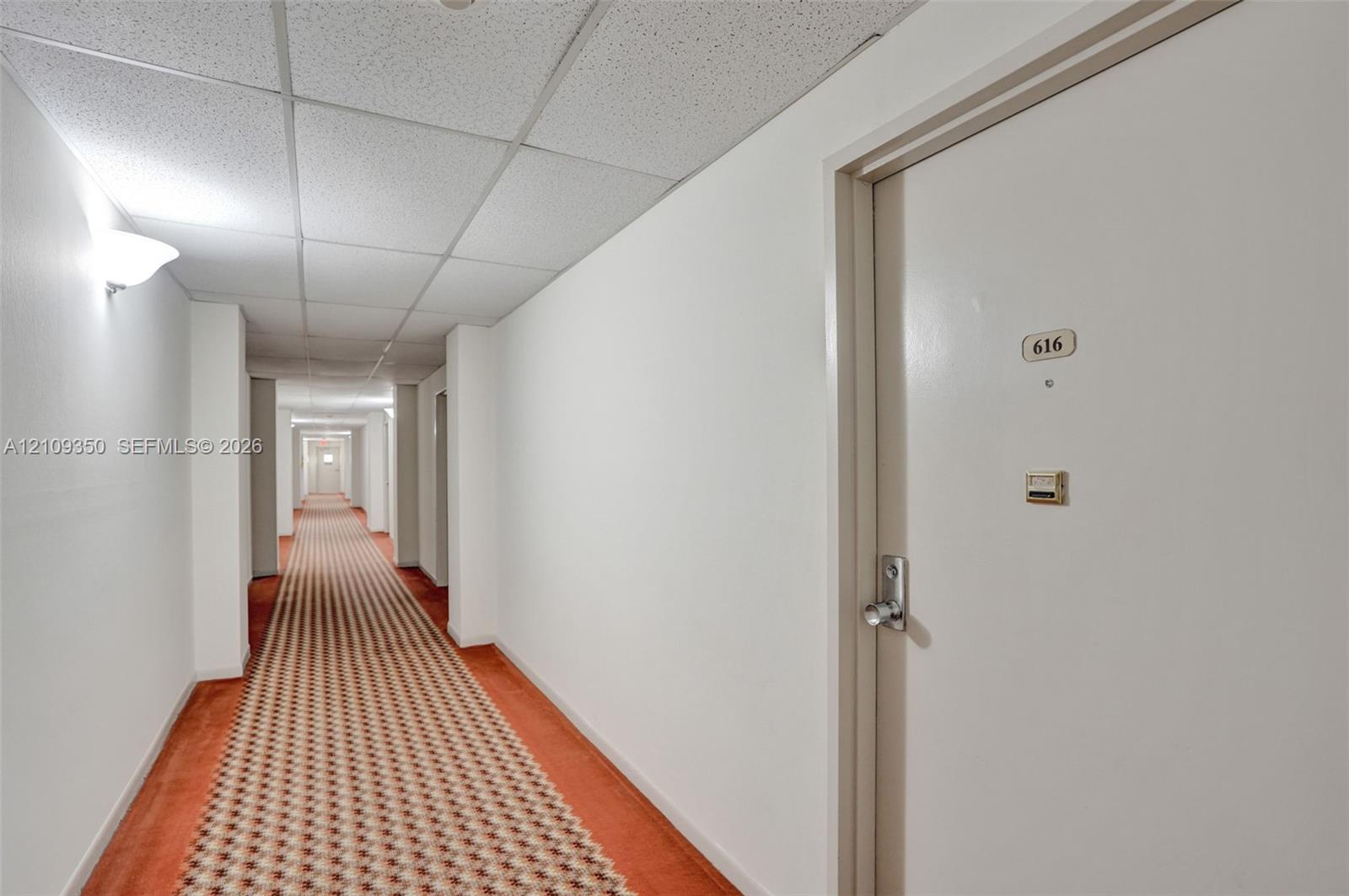 Interior hallway featuring a drop ceiling, patterned carpeting, wall-mounted lighting, and a metal-finished door handle