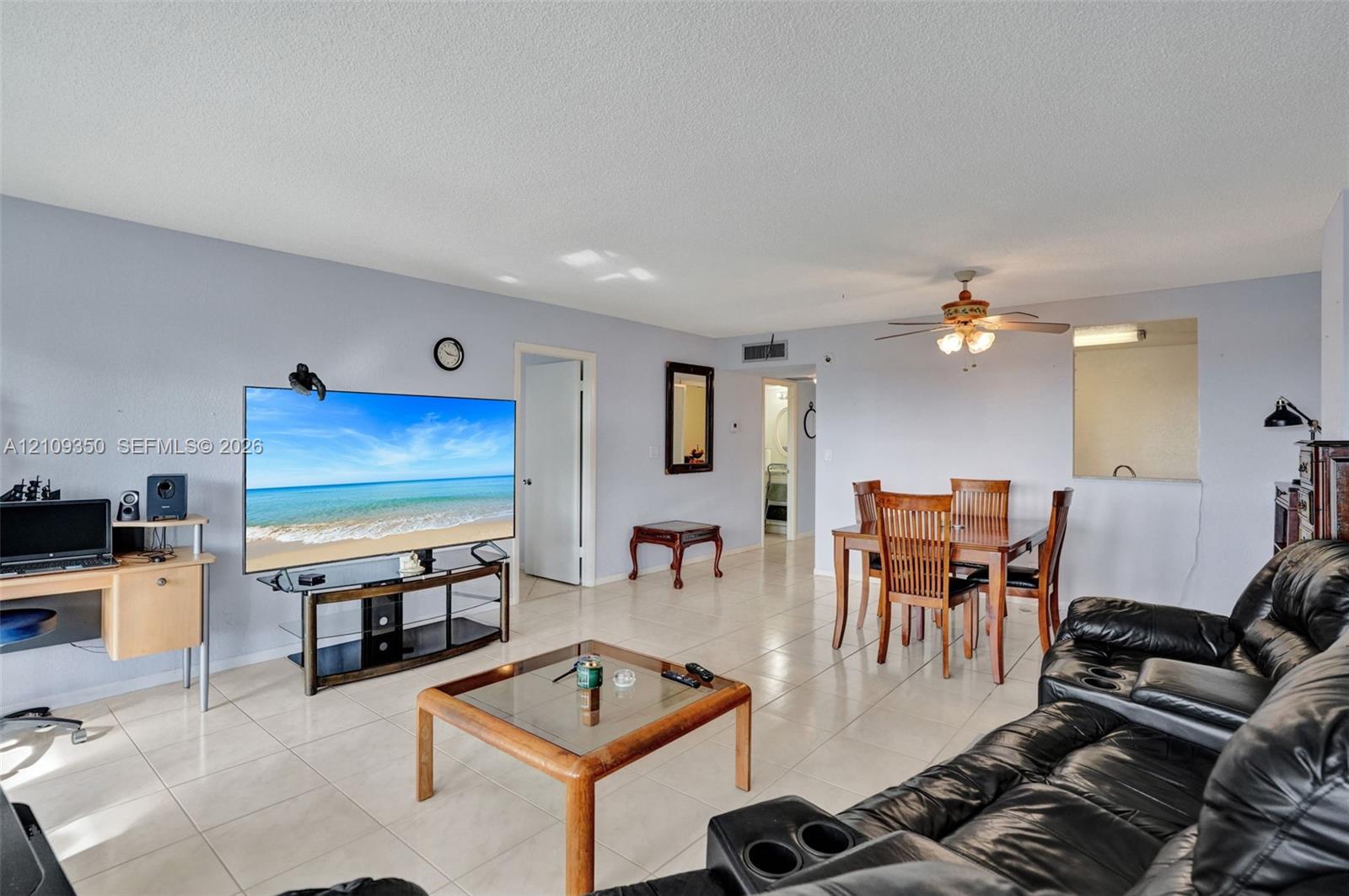 Living area featuring neutral tile flooring, a central ceiling fan with integrated lighting, and a pass-through opening to the kitchen