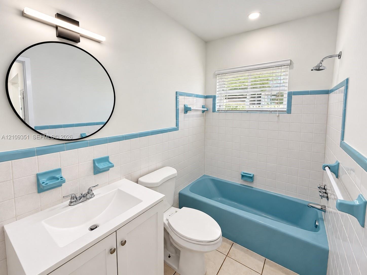 Bathroom featuring a white vanity cabinet with an integrated basin, a round wall-mounted mirror, a blue-toned soaking tub with showerhead, and white wall tile with coordinating blue trim