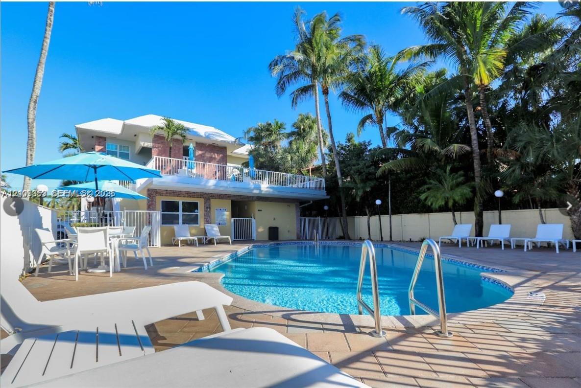 In-ground swimming pool with metal ladder, expansive paver patio, second-story railed balcony, and mature palm trees