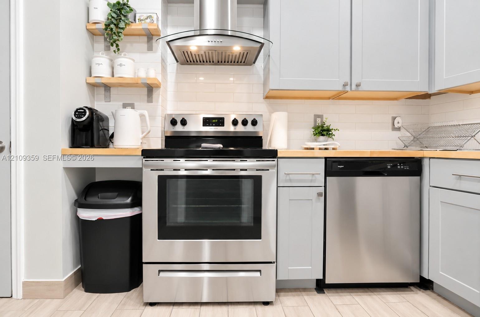 Kitchen featuring light-toned cabinetry, wood countertops, stainless steel appliances, a subway tile backsplash, and wall-mounted shelving Kitchen featuring light-toned cabinetry, wood countertops, stainless steel appliances, a subway tile backsplash, and wall-mounted shelving