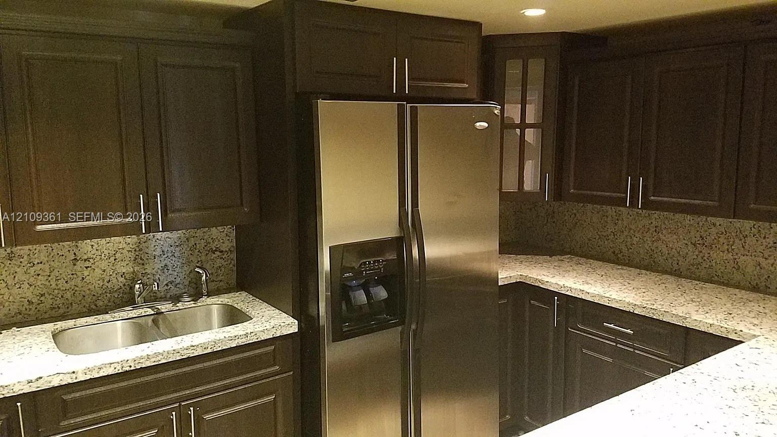 Dark wood cabinetry featuring brushed metal hardware, speckled stone countertops, stainless steel side-by-side refrigerator, under-mounted double-basin sink, and glass-paneled upper cabinet Dark wood cabinetry featuring brushed metal hardware, speckled stone countertops, stainless steel side-by-side refrigerator, under-mounted double-basin sink, and glass-paneled upper cabinet