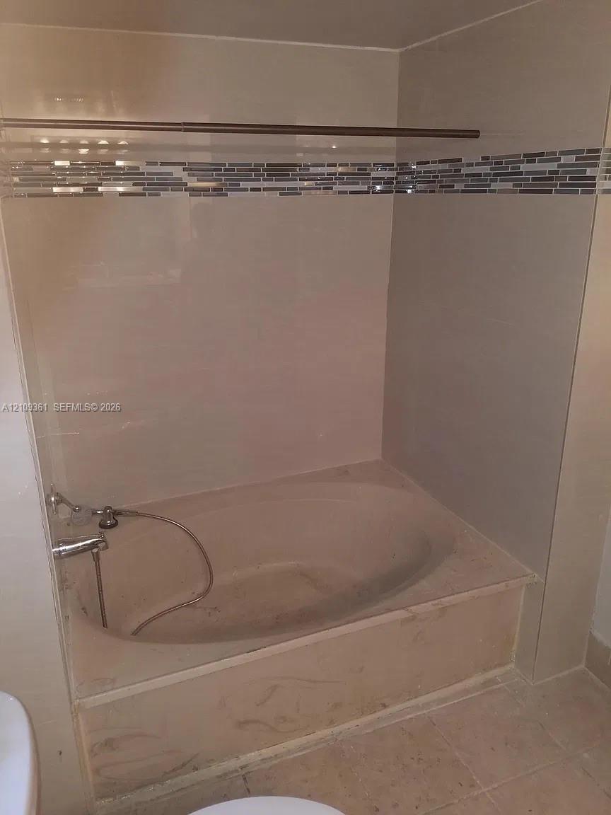 Built-in alcove soaking tub featuring a tiled surround, integrated stone-finish deck, mosaic glass accent tiling, and a chrome hand-held shower fixture Built-in alcove soaking tub featuring a tiled surround, integrated stone-finish deck, mosaic glass accent tiling, and a chrome hand-held shower fixture