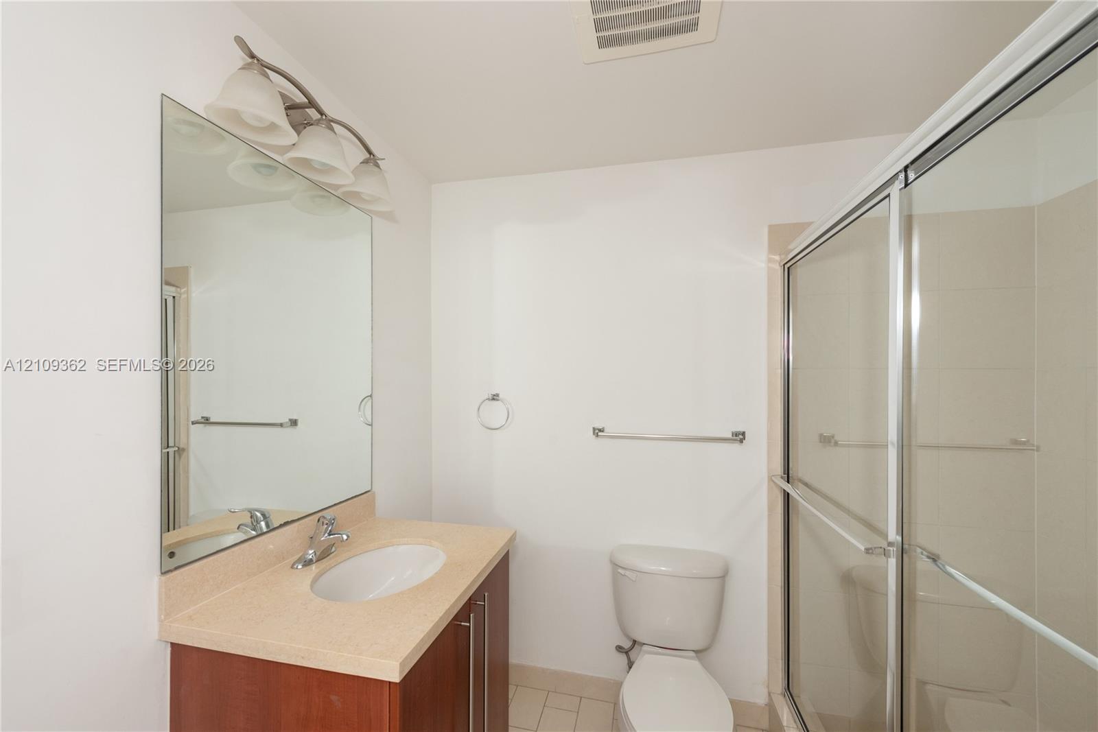 Bathroom featuring a single-sink vanity with a solid-surface countertop, full-height wall mirror, multi-bulb light fixture, and a glass-enclosed shower stall