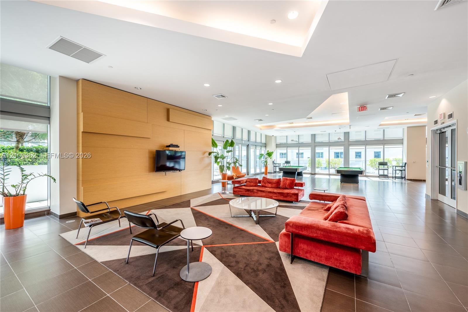 Spacious common area featuring large-format tile flooring, recessed ceiling lighting, tray ceiling details, floor-to-ceiling windows, and wood-paneled accent wall